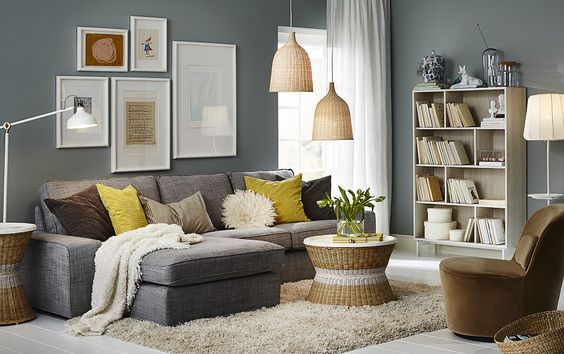 A living room furnished with a gray two-seat sofa combined with a chaise lounge. Shown together with a round tray table and an easy chair that swivels, covered with dark beige velvet.